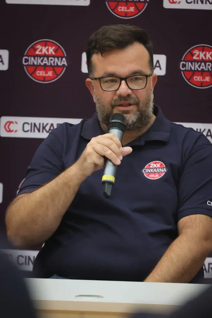 Član strokovnega štaba z mikrofonom pred klubskim panojem.