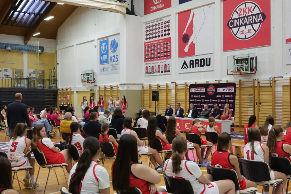 Košarkarice in gostje spremljajo novinarsko konferenco v dvorani.